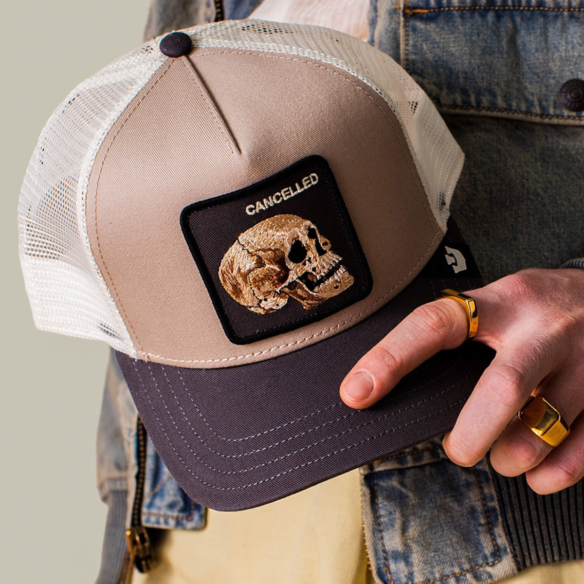 A hand holds "The Cancelled Skull" trucker hat by Goorin Bros.—a beige and black cap with a stitched skull patch reading CANCELLED. Paired with gold rings and a denim jacket, it's a bold way to speak your mind.