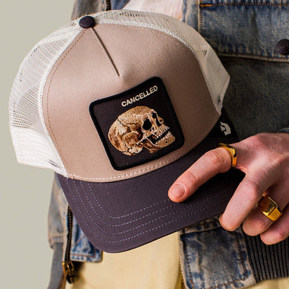 A hand holds "The Cancelled Skull" trucker hat by Goorin Bros.—a beige and black cap with a stitched skull patch reading CANCELLED. Paired with gold rings and a denim jacket, it's a bold way to speak your mind.