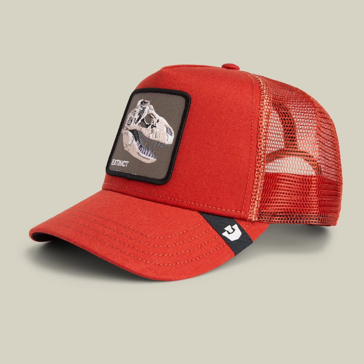 The Extinct Skull by Goorin Bros is a red mesh trucker hat featuring a dinosaur skull embroidered patch on the front and a small logo tab on the bill, displayed against a neutral background.