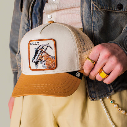 A person holds the Goat Trucker hat by Goorin Bros.—a beige and brown cap with a bold embroidered goat patch labeled "G.O.A.T." They're accessorized with gold rings, a denim jacket, light pants, and pearl accents in neutral tones.