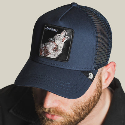 A man wears the Goorin Bros. Lone Wolf Trucker cap—a navy blue mesh-back hat featuring an embroidered howling wolf patch and Lone Wolf text, giving off bold outlaw vibes.