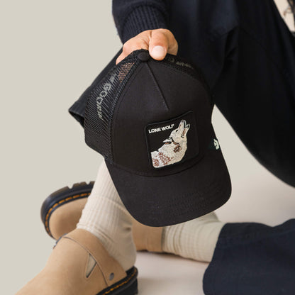 A person wears the Goorin Bros. Lone Wolf Trucker, a black mesh cap with a patch showing a wolf and "Lone Wolf" text, paired with beige socks, sandals, and dark pants for an effortlessly cool, rebellious style.