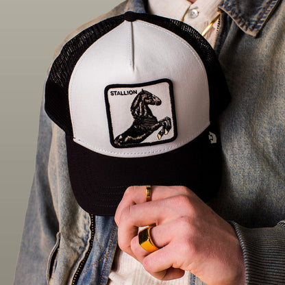Someone wearing a denim jacket and gold rings holds the Goorin Bros. Stallion Trucker, a black and white trucker hat featuring a stallion patch.