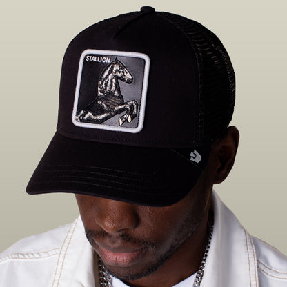 A person wearing the Goorin Bros. Stallion Trucker hat—black and white with a horse patch—paired with a white jacket and a silver chain necklace.