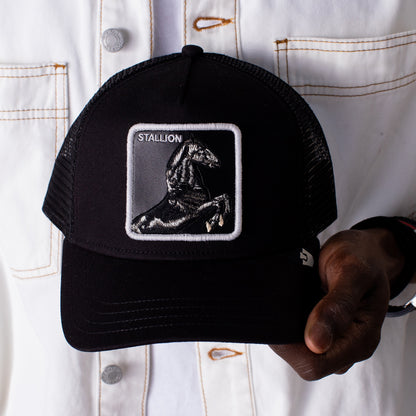 A person in a white jacket holds the Stallion Trucker hat by Goorin Bros., featuring a black and white design with a patch showing an embroidered black horse and the word "Stallion.