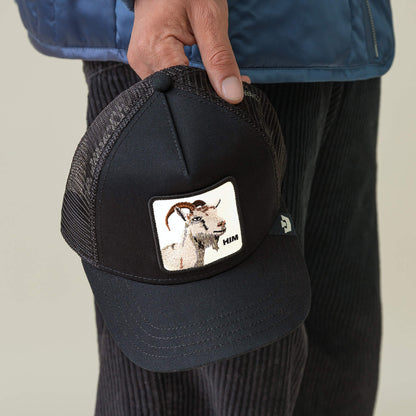 A person holds the Him Trucker cap by Goorin Bros., a black mesh hat with a front patch featuring an embroidered GOAT and the word "HIM.