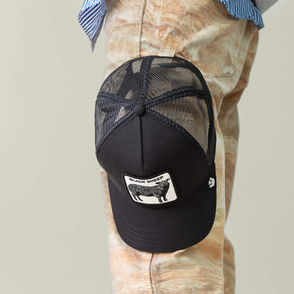 The Goorin Bros. Black Sheep Trucker cap hangs from a person’s knee, paired with beige marbled pants and a blue-striped shirt—showcasing an effortlessly bold style.
