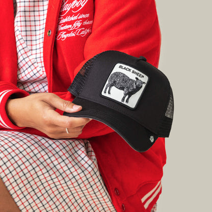A person in a red jacket and checkered dress holds the Goorin Bros. Black Sheep Trucker hat, featuring a bold patch with a black sheep and the words BLACK SHEEP—ideal for anyone who embraces individuality.