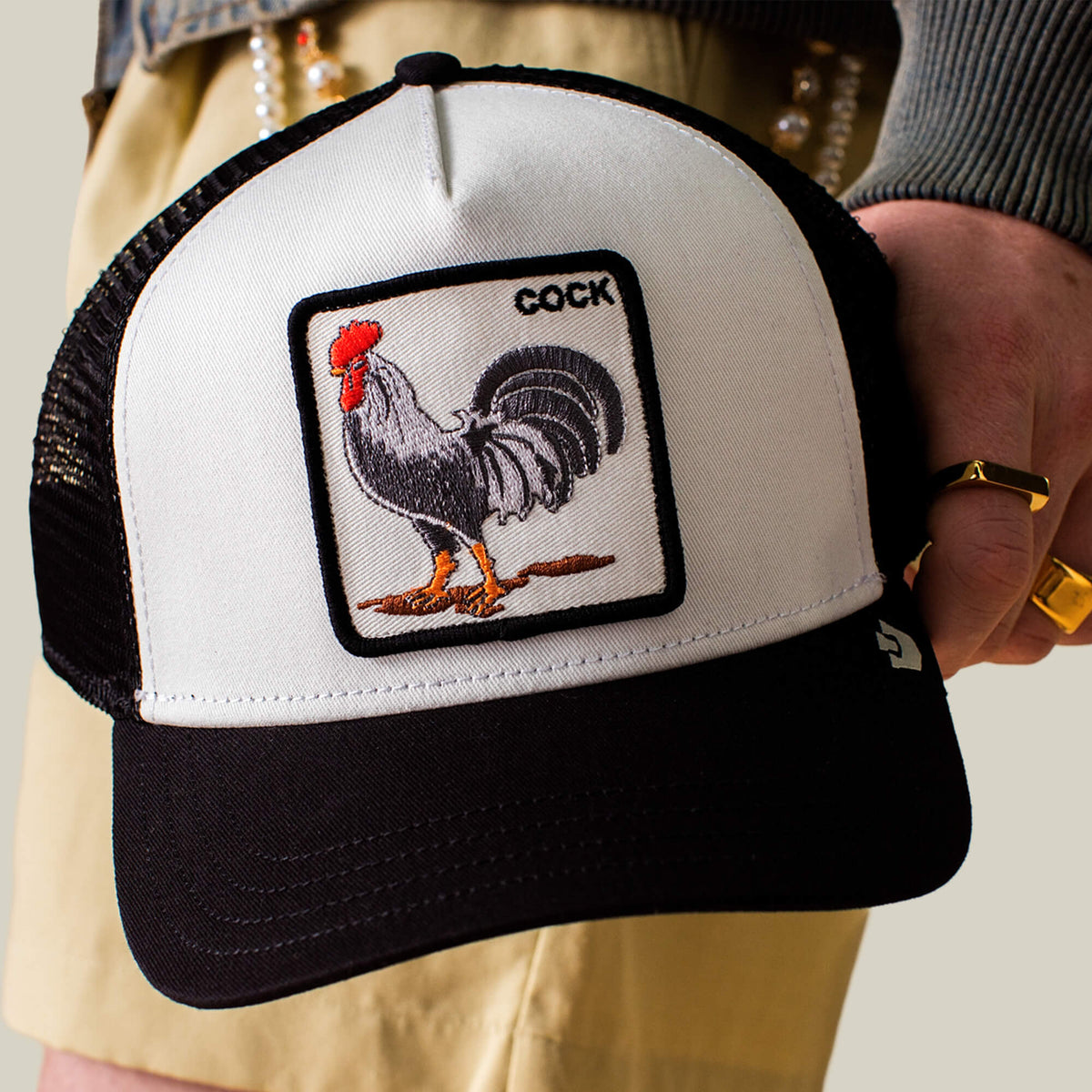 A person holds a Goorin Bros. Rooster Trucker hat in black and white, featuring a bold rooster patch with the word "COCK" on it.