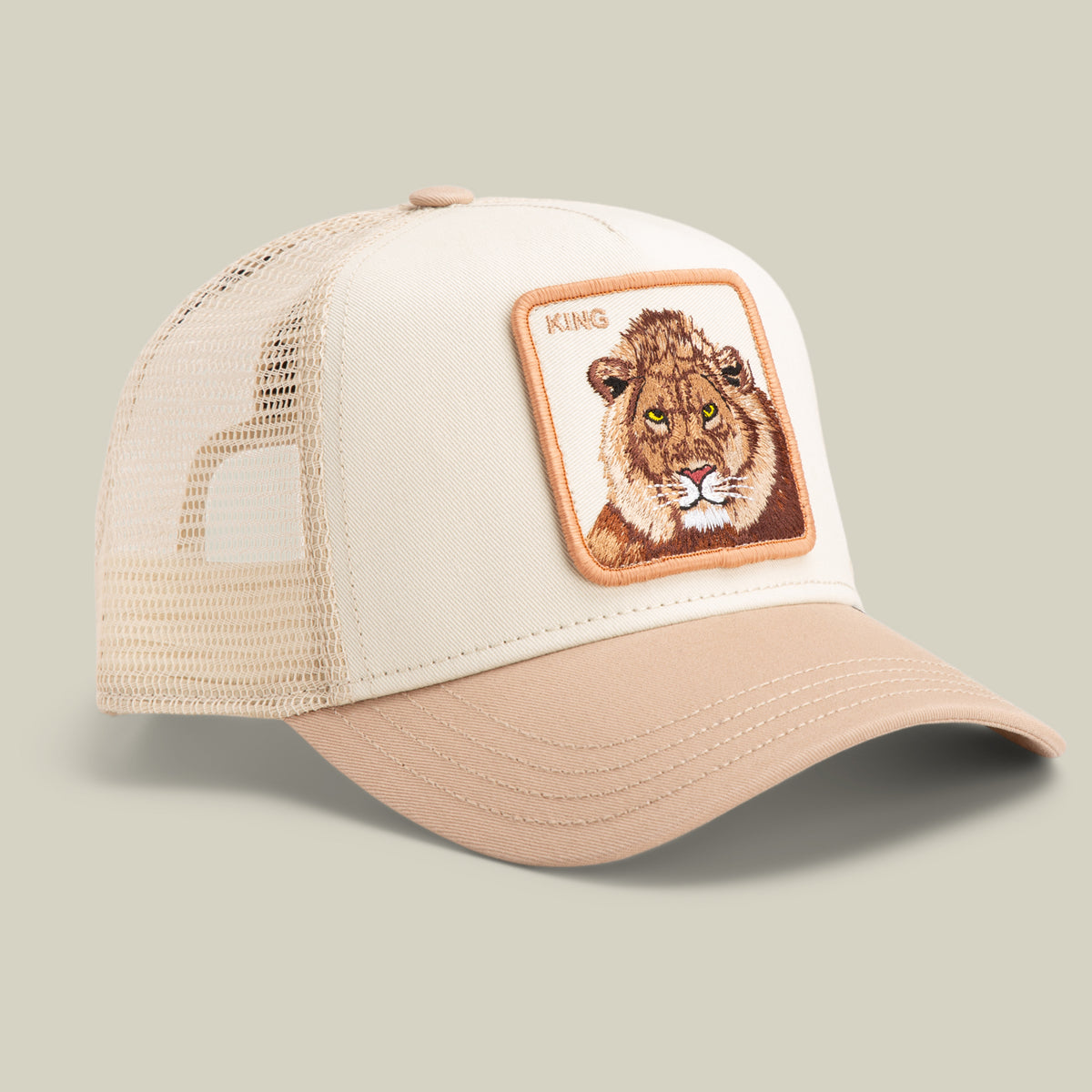 The Goorin Bros. King Trucker cap features beige and tan panels, a mesh back, and a front patch with an embroidered lion head and "KING" text.