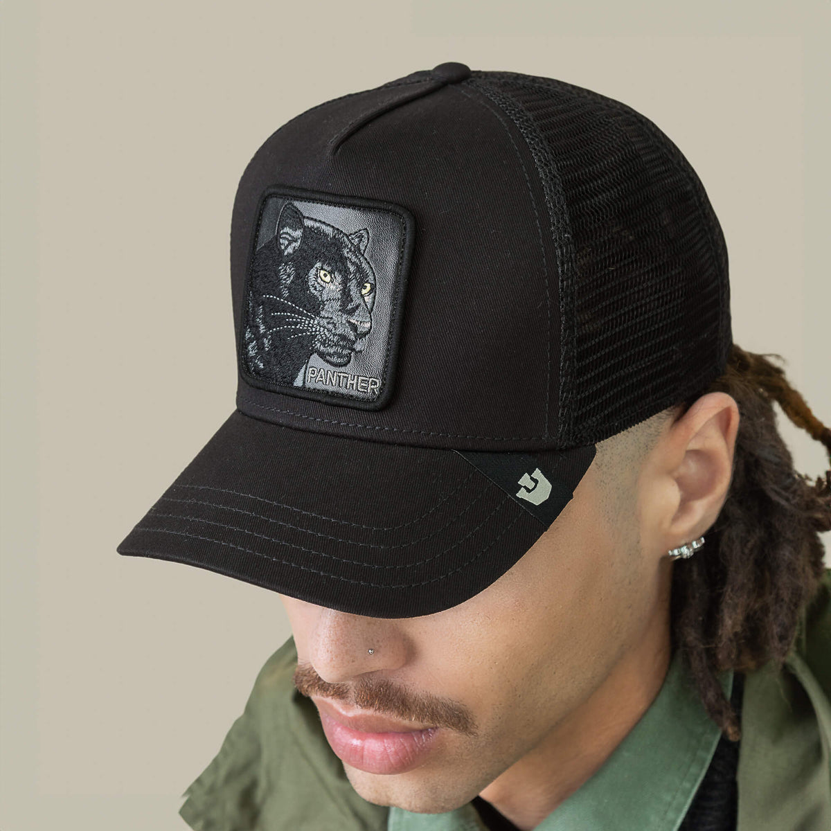 A person sports the Goorin Bros. Panther Trucker—a black mesh baseball cap featuring a stitched panther patch—paired with a green collared shirt and jacket.