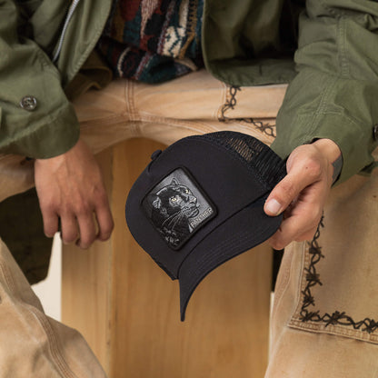 A person holds the Goorin Bros. Panther Trucker hat, featuring a black cat patch, while sporting an olive jacket and light-colored pants with decorative stitching.