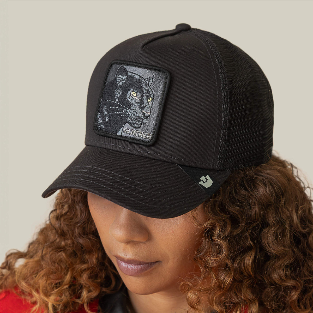 A woman with curly hair wears the Goorin Bros. Panther Trucker—a stylish black mesh trucker cap featuring a rectangular patch with an embroidered black panther design on the front.
