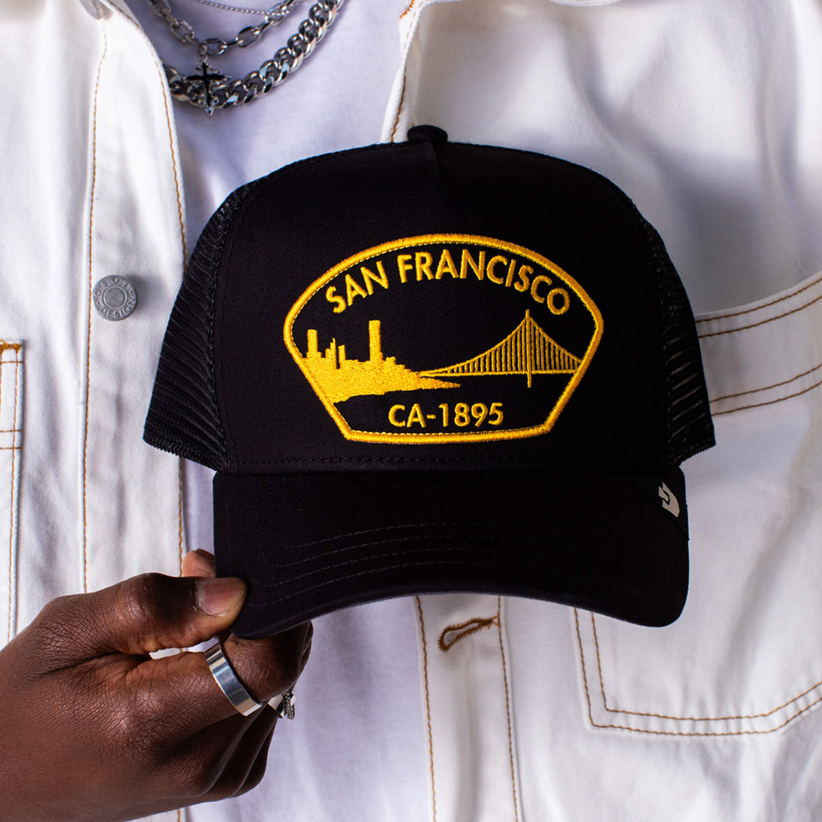 A person in a white jacket holds a black Goorin Bros. San Francisco hat with a yellow patch featuring the city skyline and Golden Gate Bridge.