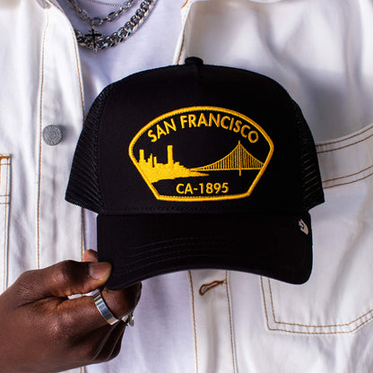 A person in a white jacket holds a black Goorin Bros. San Francisco hat with a yellow patch featuring the city skyline and Golden Gate Bridge.