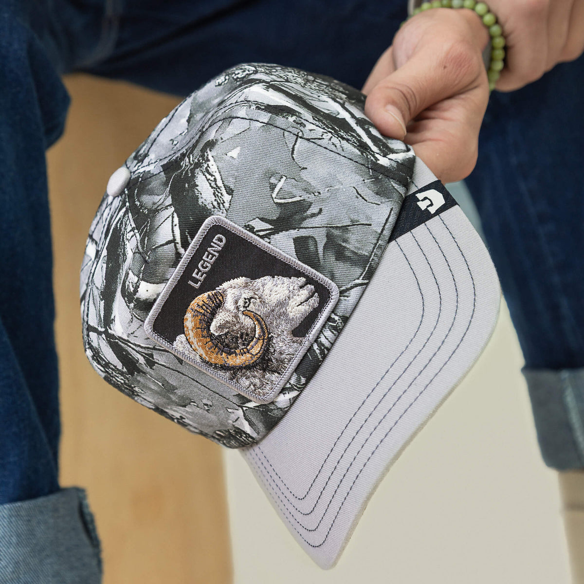 Someone holds the Legend Camo baseball cap by Goorin Bros.—a gray frosted camo hat with a bighorn patch labeled "Legend" on the front, white brim, and Low Pro Fit.