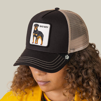 A person wears the Bad Boy Trucker by Goorin Bros., a black and tan mesh cap featuring a Rottweiler patch with "BAD BOY" on the front, adding an edgy statement as they look down, face partially visible.