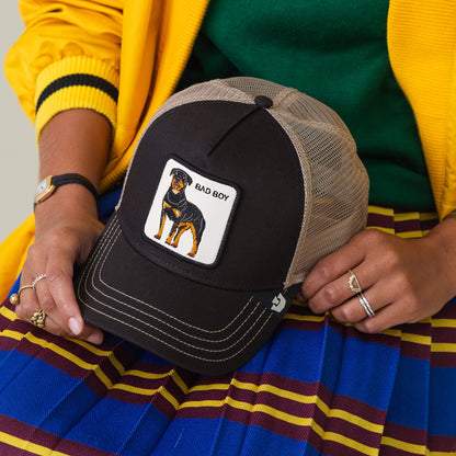 A person sports the Goorin Bros. Bad Boy Trucker—a beige and black mesh hat with a Rottweiler patch—paired with a yellow jacket, green top, and blue skirt decorated with red and yellow stripes for an edgy look.