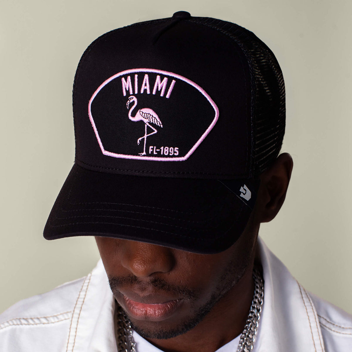 A person sports the Goorin Bros. "Miami" black mesh cap with a pink flamingo and MIAMI FL-1895 text, paired with a white jacket and silver jewelry, all together reflecting Miami’s vibrant energy.