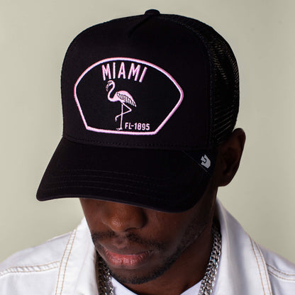 A person sports the Goorin Bros. "Miami" black mesh cap with a pink flamingo and MIAMI FL-1895 text, paired with a white jacket and silver jewelry, all together reflecting Miami’s vibrant energy.