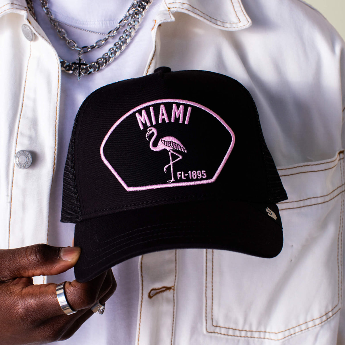 A person in a white jacket holds the Goorin Bros. "Miami" black trucker hat, featuring a pink flamingo and "Miami FL-1895" embroidery on the front, embodying South Beach's playful flair.