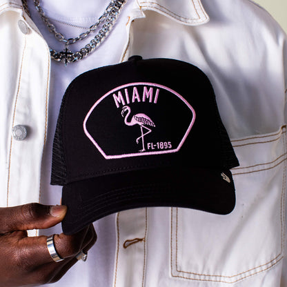 A person in a white jacket holds the Goorin Bros. "Miami" black trucker hat, featuring a pink flamingo and "Miami FL-1895" embroidery on the front, embodying South Beach's playful flair.