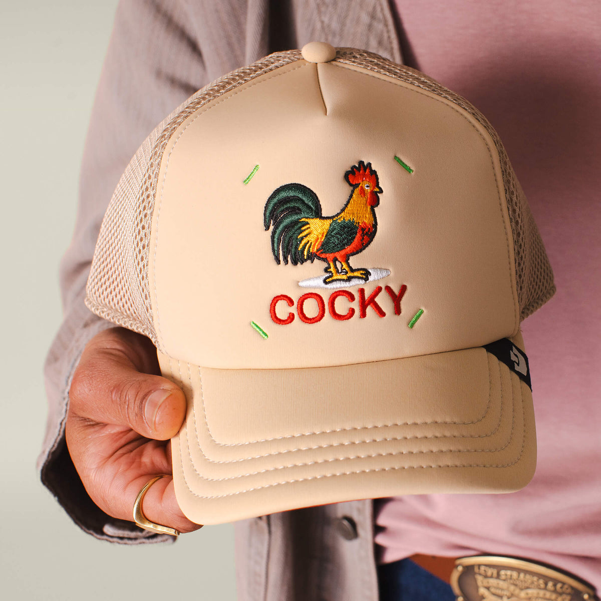A person holds the Neo Farm - Cocky Rooster trucker hat by Goorin Bros., featuring neutral beige tones and a rooster embroidery above bold red "COCKY" lettering.