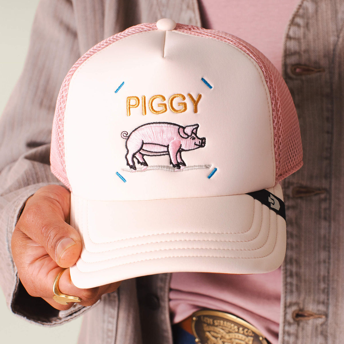 Someone holds the Neo Farm - Piggy Pig hat by Goorin Bros., a pink cap with breathable mesh, "PIGGY" stitched on the front, and an embroidered pig.