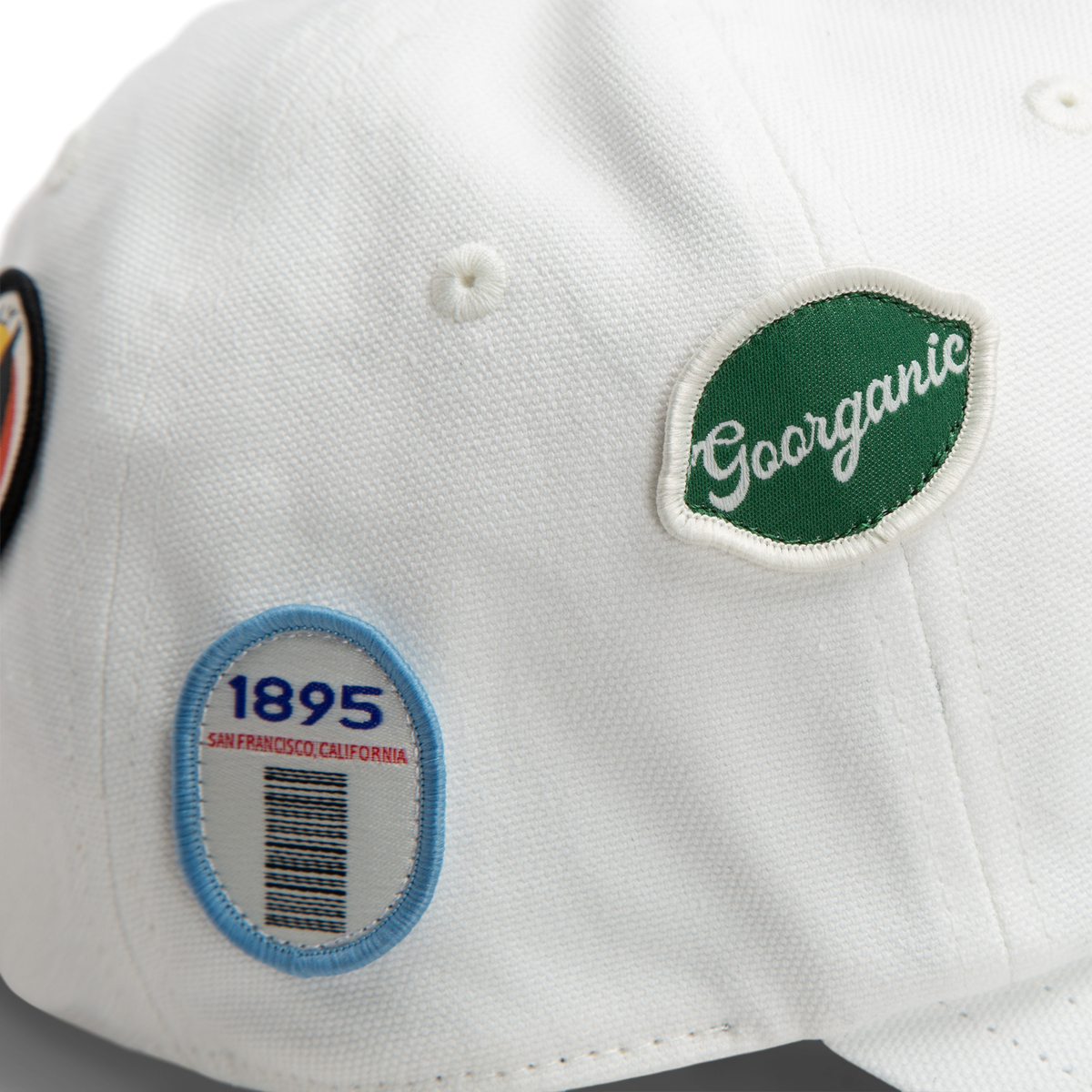 Close-up of a white Pop Appeal cap by Goorin Bros., featuring two embroidered badges—one green with Gorganic text and another oval-shaped with "1895 SAN FRANCISCO, CALIFORNIA" and a barcode design.