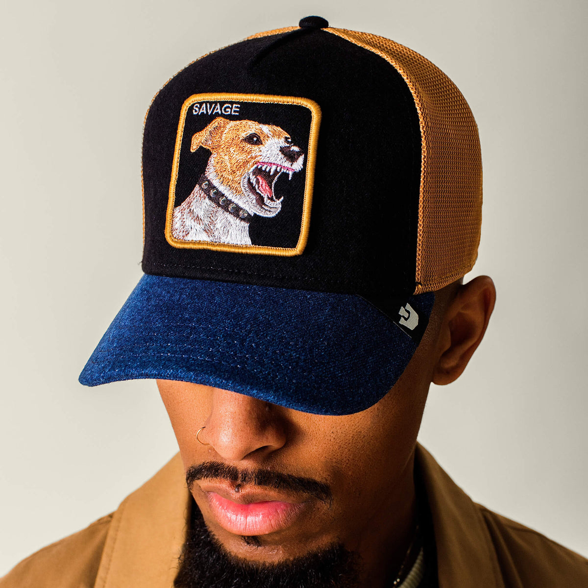 A man wearing the Goorin Bros. Savage Dawg trucker hat, featuring a mixed denim colorway, blue brim, and a snarling dog patch labeled SAVAGE on the front.