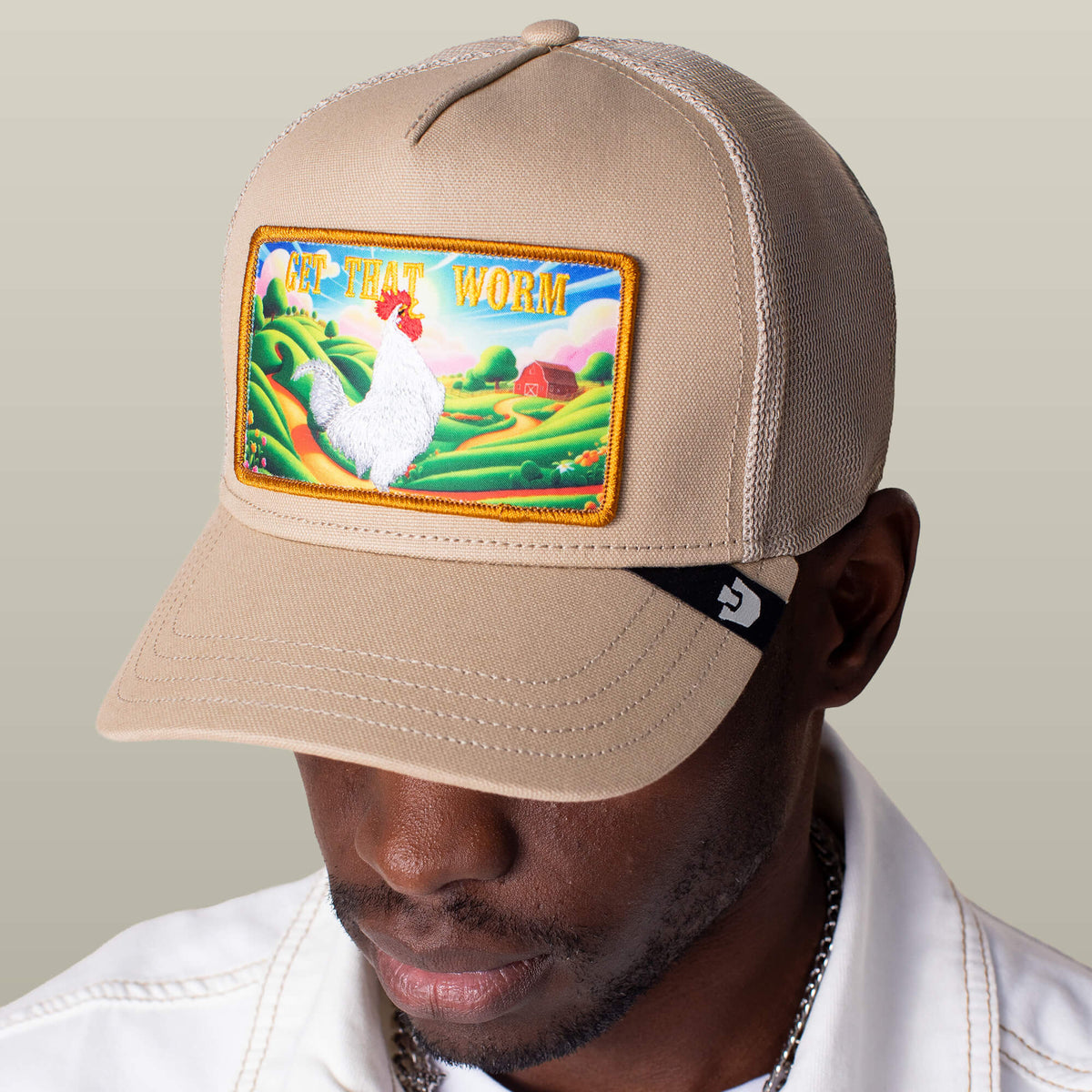 A person wears the Goorin Bros. Greener Grass - Get that Worm beige trucker hat, featuring a vibrant patch with a white chicken and colorful farm scene.