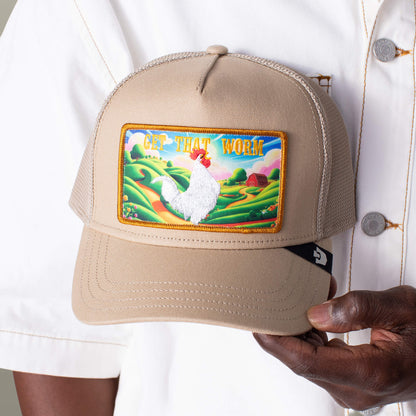 A person holds the Greener Grass - Get that Worm tan trucker hat by Goorin Bros., featuring a colorful rooster patch and "GET THAT WORM" text, against a scenic farm backdrop.