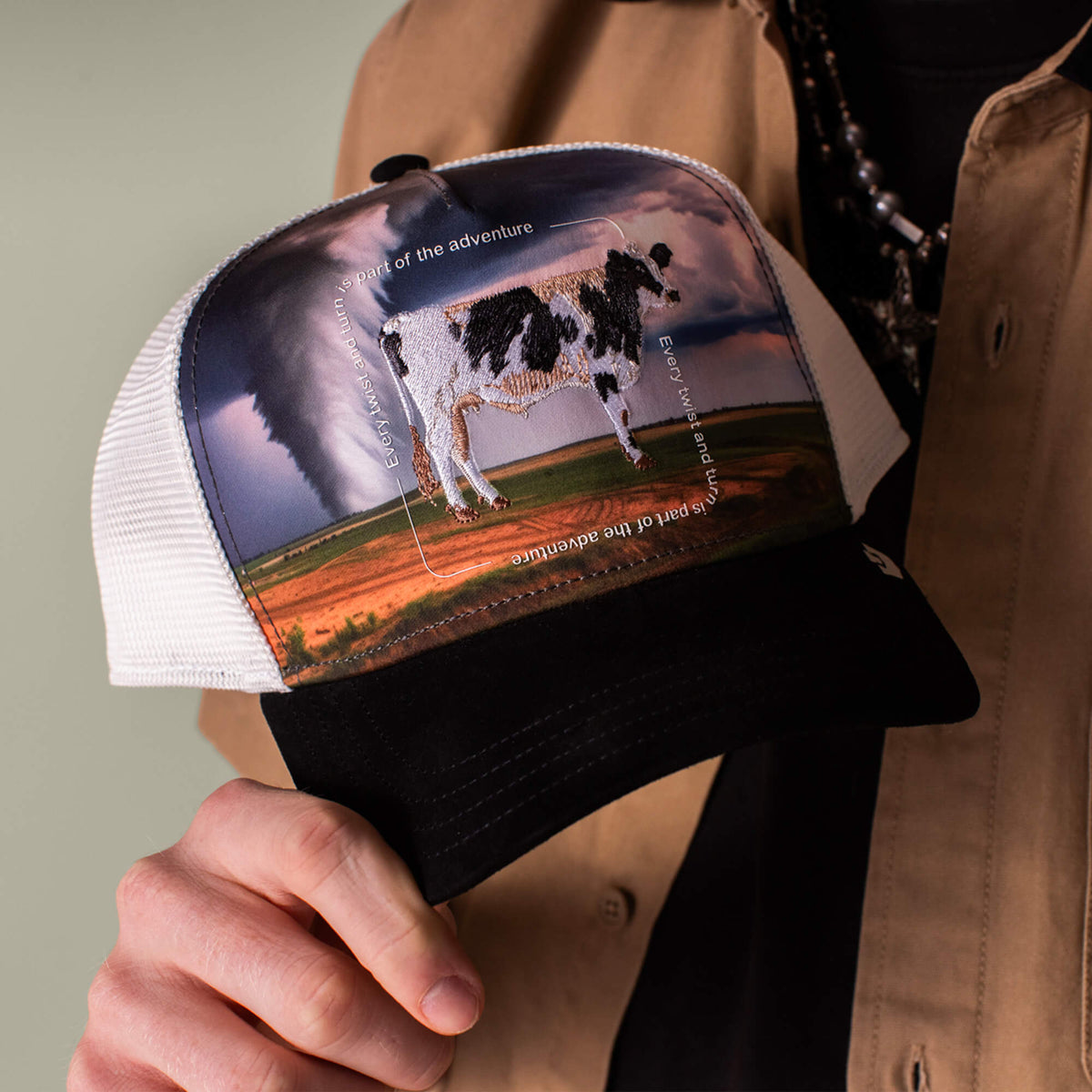 A person holds the Goorin Bros. "Through the Storm - Cow" black and white trucker hat, featuring an embroidered cow patch with circling white text and a tornado in the background, evoking a sense of adventure.