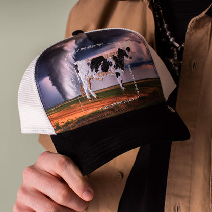A person holds the Goorin Bros. "Through the Storm - Cow" black and white trucker hat, featuring an embroidered cow patch with circling white text and a tornado in the background, evoking a sense of adventure.