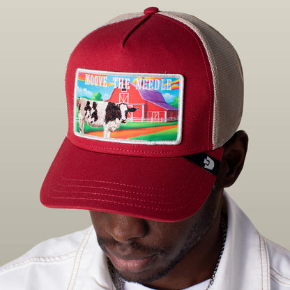 A man wears the Goorin Bros. Greener Grass - Moove the Needle trucker hat in red and beige, featuring a farm patch with a cow, barn, and "Moove The Needle" text.