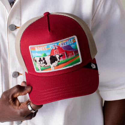 A person holds the Greener Grass - Moove the Needle red and beige trucker hat by Goorin Bros., featuring a farm patch with a cow, barn, field, and the words “MOOVE THE NEEDLE.”.