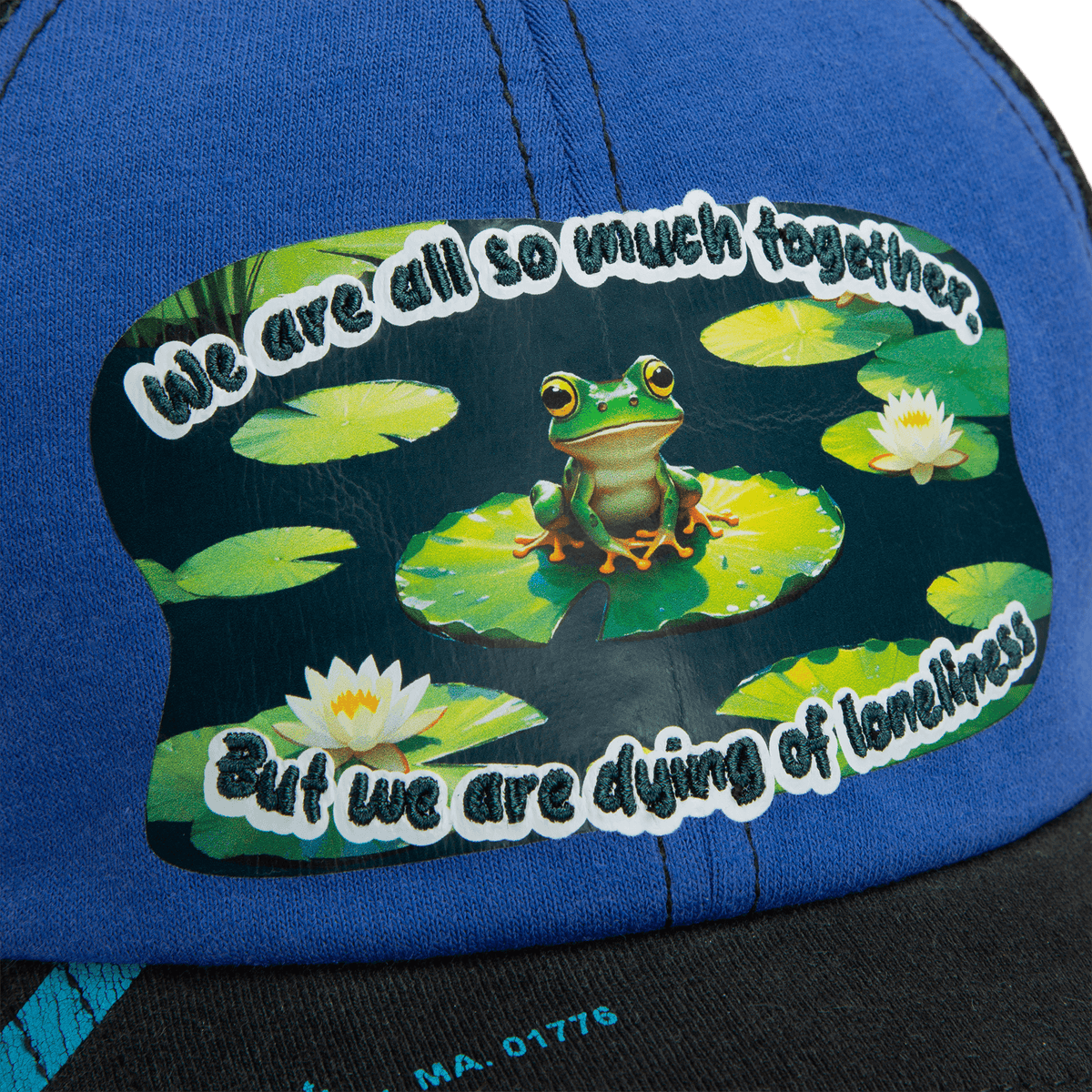A sticker on the blue Lil Sohei cap by Goorin Bros. shows a frog on a lily pad among water lilies with the text: We are all so much together. But we are dying of loneliness. Explore unique hats that tell your story.