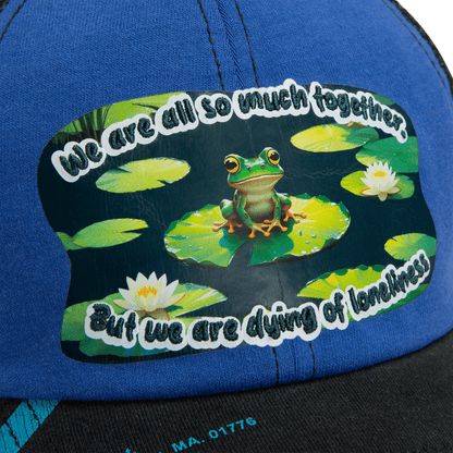 A sticker on the blue Lil Sohei cap by Goorin Bros. shows a frog on a lily pad among water lilies with the text: We are all so much together. But we are dying of loneliness. Explore unique hats that tell your story.
