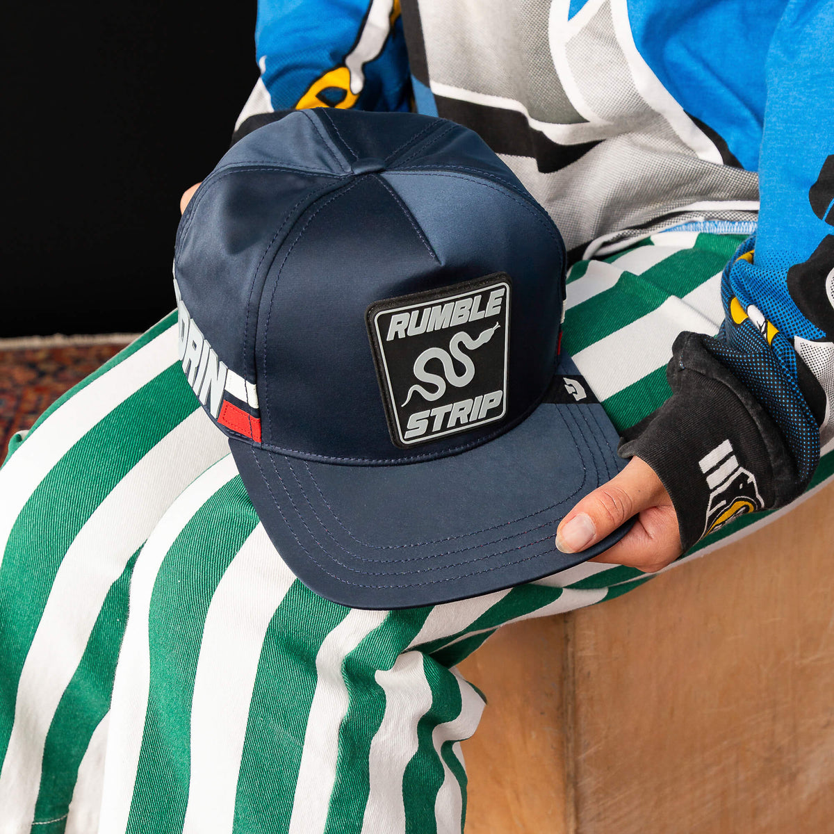A person in green and white striped pants and a colorful top holds the Goorin Bros. Pole Position - Rumble Strip cap—navy blue with a Rumble Strip patch, ideal for drift drivers seeking standout style.