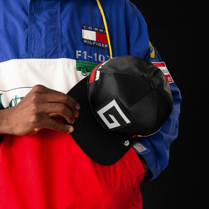 A person in a color-block Tommy Hilfiger jacket holds the Goorin Bros. Pole Position - Road Kill black racing cap featuring a geometric G logo and chrome satin stripes on the front, against a black background.