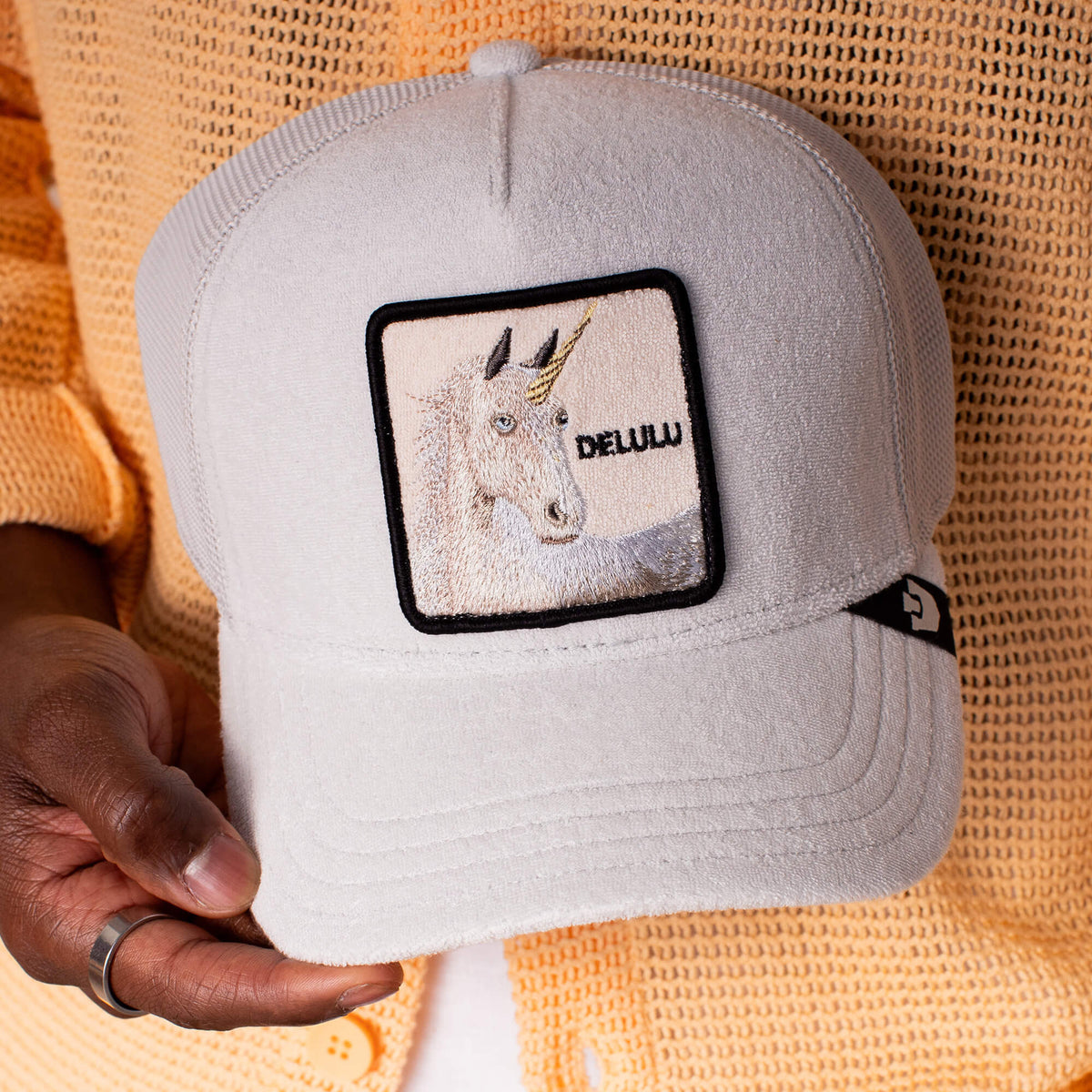 A person holds the Goorin Bros. Terry Cloth Unicorn cap, made from soft light gray fabric and featuring a patch with a unicorn and "DELULU" embroidered on it.