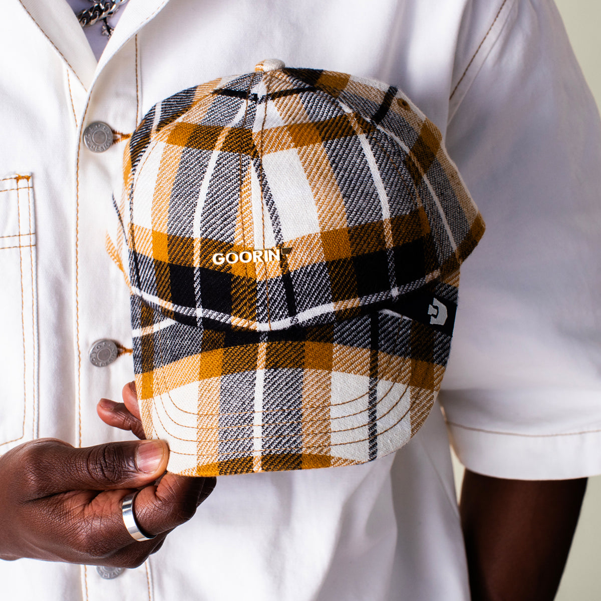 A person in a white jacket holds the Bespoke Wordmark V2 cap by Goorin Bros., featuring a yellow, black, and white plaid design with the GOORIN wordmark.