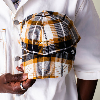 A person in a white jacket holds the Bespoke Wordmark V2 cap by Goorin Bros., featuring a yellow, black, and white plaid design with the GOORIN wordmark.