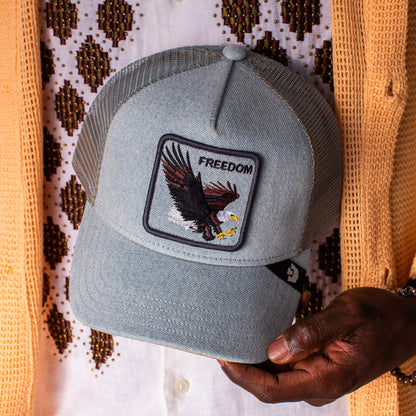A person holds the Goorin Bros. Denim Eagle—a light blue denim trucker hat with an eagle patch and "FREEDOM" embroidered on the front.