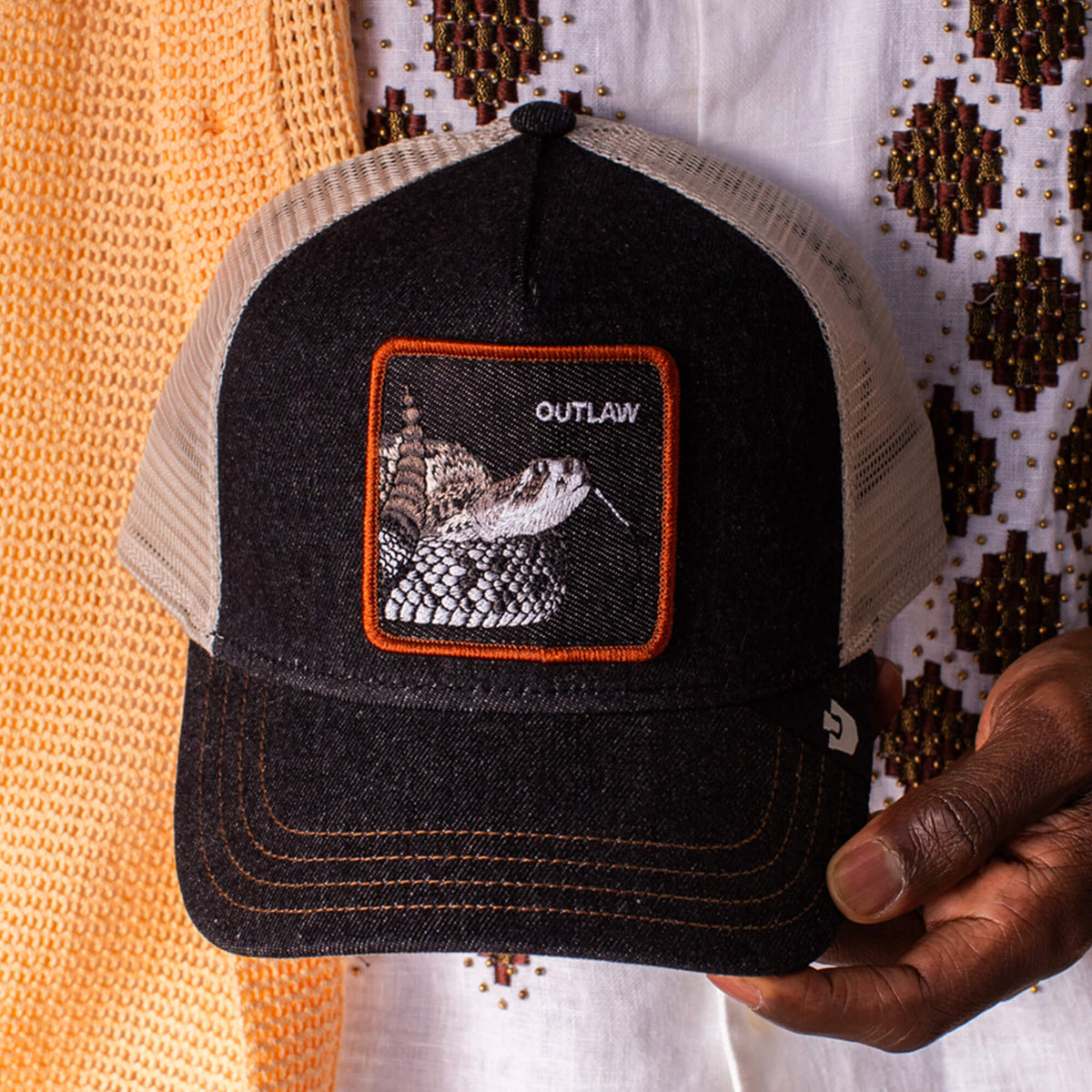 A person holds a Goorin Bros. Denim Snake trucker hat in black and beige, featuring a rattlesnake patch on the front.