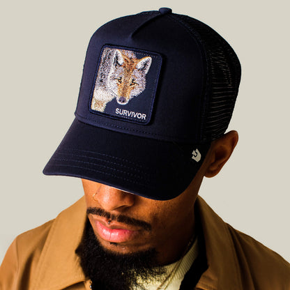 A man wears the Goorin Bros. Mirage Survivor black mesh baseball cap featuring a fox patch labeled “SURVIVOR,” paired with a brown jacket and looking down against a plain beige background, capturing the rugged appeal of this signature hat.