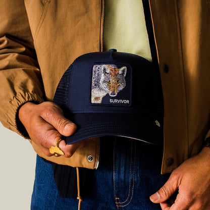 A person in a brown jacket holds the Mirage Survivor navy trucker hat by Goorin Bros., featuring a patch with a fox and the word SURVIVOR.