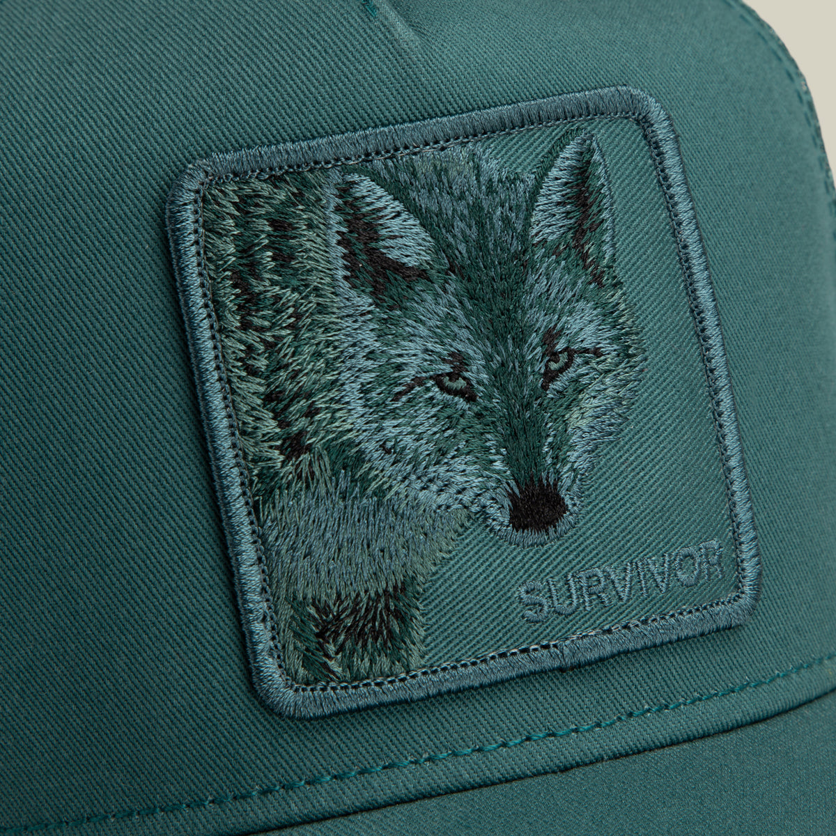 Goorin Bros. Mirage Survivor teal trucker hat featuring a square patch embroidered with a wolf’s face and the word SURVIVOR.