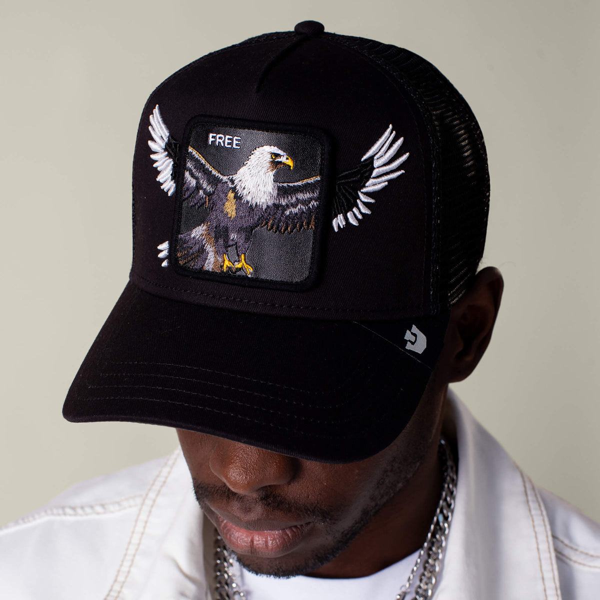 A person wearing the Goorin Bros. Wings Free black eagle cap with mesh back and "FREE" embroidered on the front, paired with a white jacket and silver chain.