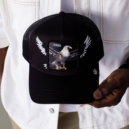 A person holds the Goorin Bros. Wings Free hat, a classic black trucker cap with breathable mesh back, featuring an embroidered eagle with outstretched wings and "FREE" above it.